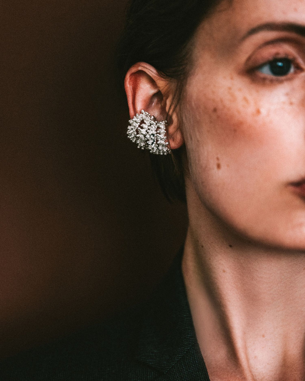 A close-up of a model's ear wearing a sparkling silver cluster ear cuff, set against a dark brown background.