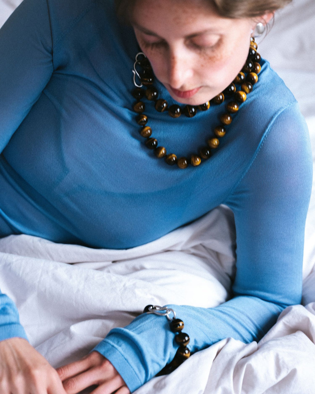 A woman wearing a bright blue long-sleeve top and layered brown tiger-eye beaded bracelets and necklace, sitting on white bedding.