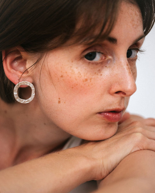 A close-up portrait of a woman wearing a textured silver hoop earring with a clear pavé-like setting. The photo focuses on her ear and profile against a soft light gray background.