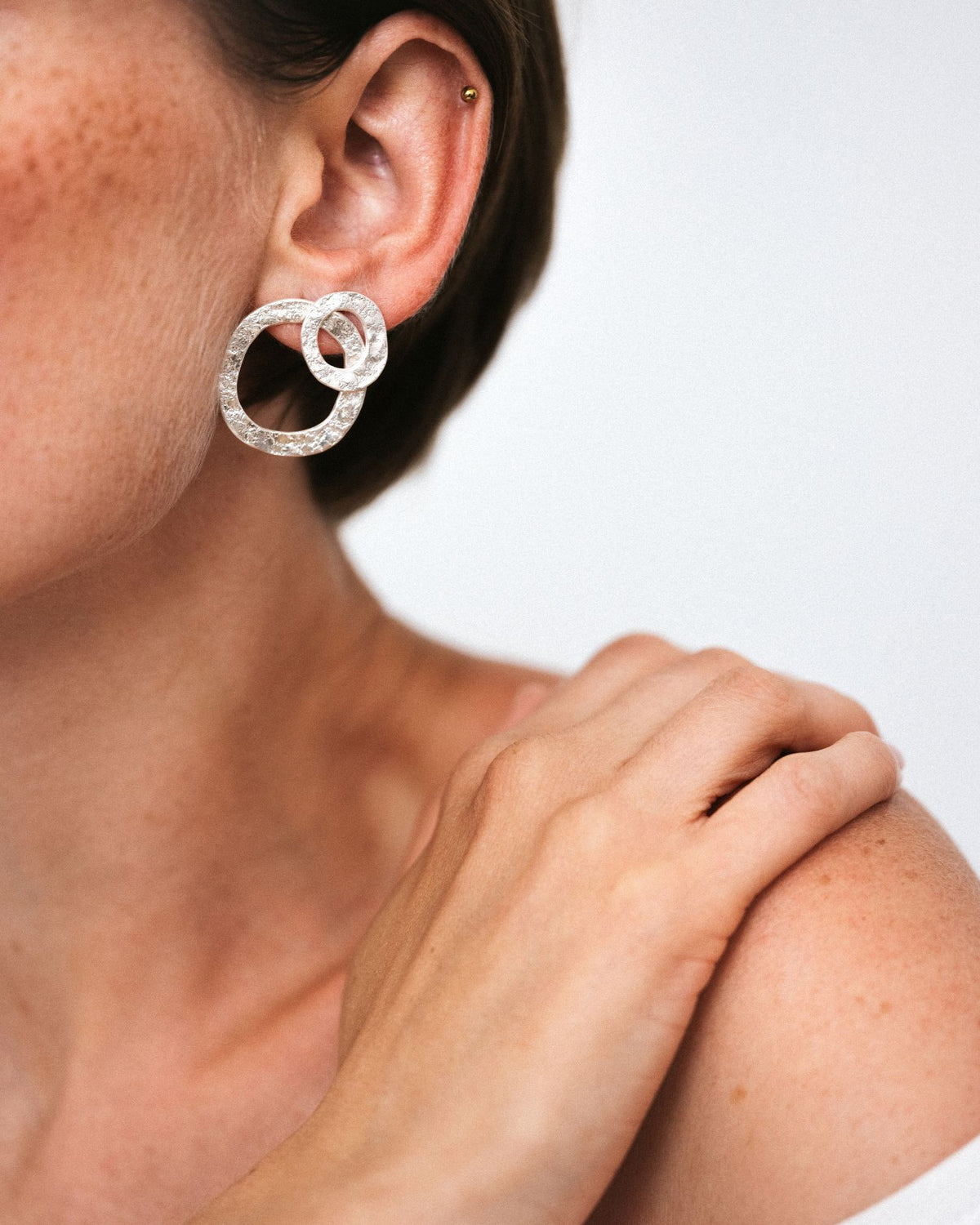 A close-up of a model's ear wearing a pair of silver interlocking circular hoop earrings with textured pavé detailing, against a clean light gray background.