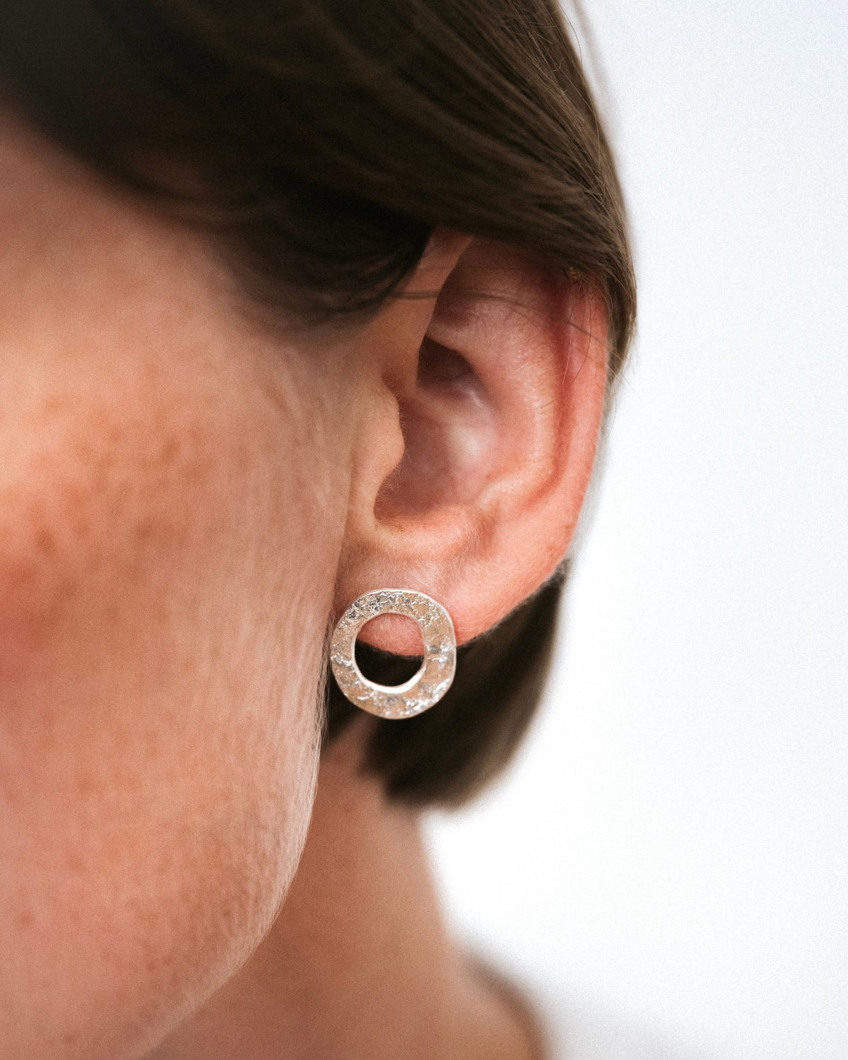 A close-up of a person's ear wearing a small silver hoop earring with a textured, glittery outer ring and a central cutout.
