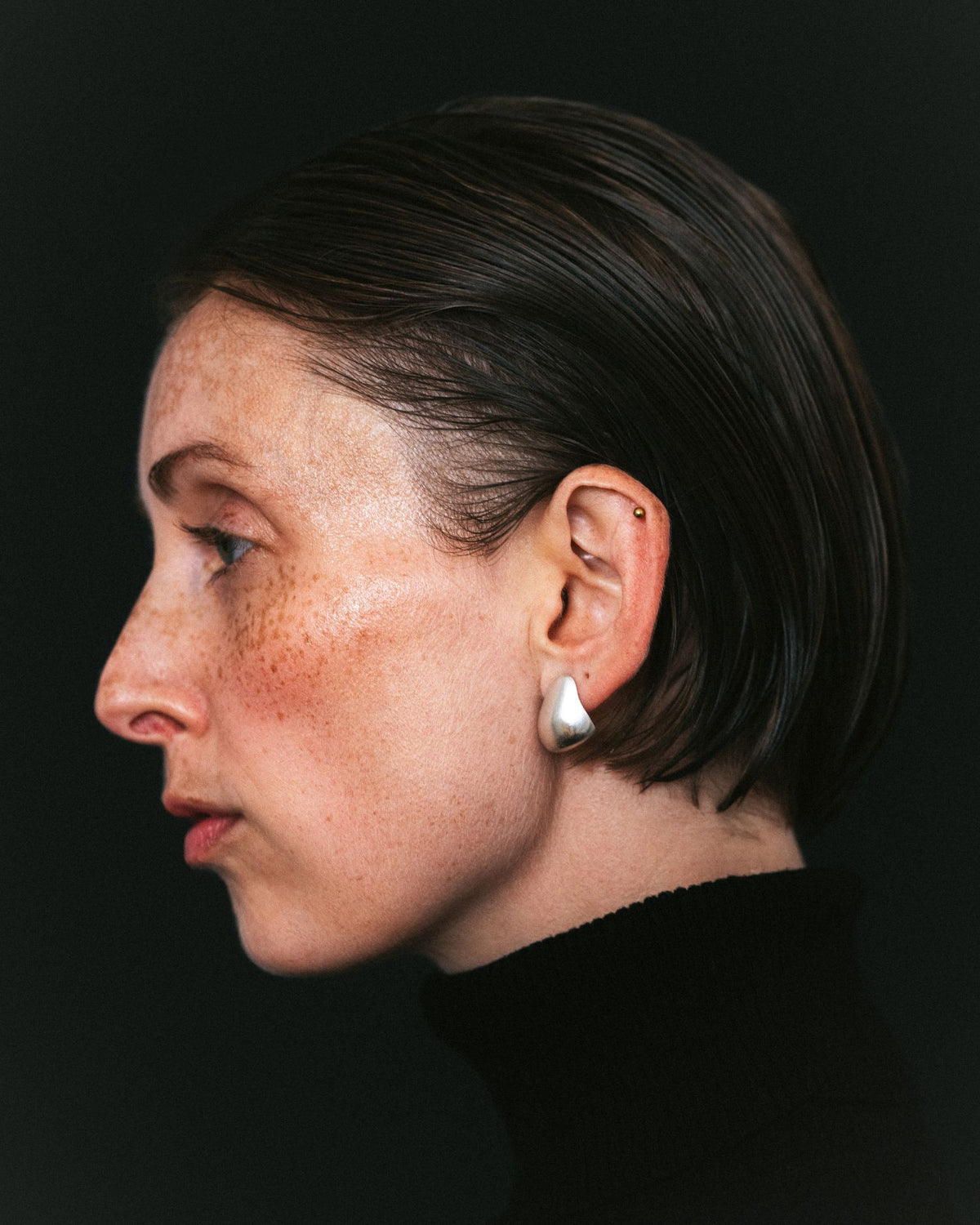 A close-up side profile portrait of a woman with short dark hair, freckles, and a small silver teardrop-shaped stud earring, set against a solid black background.