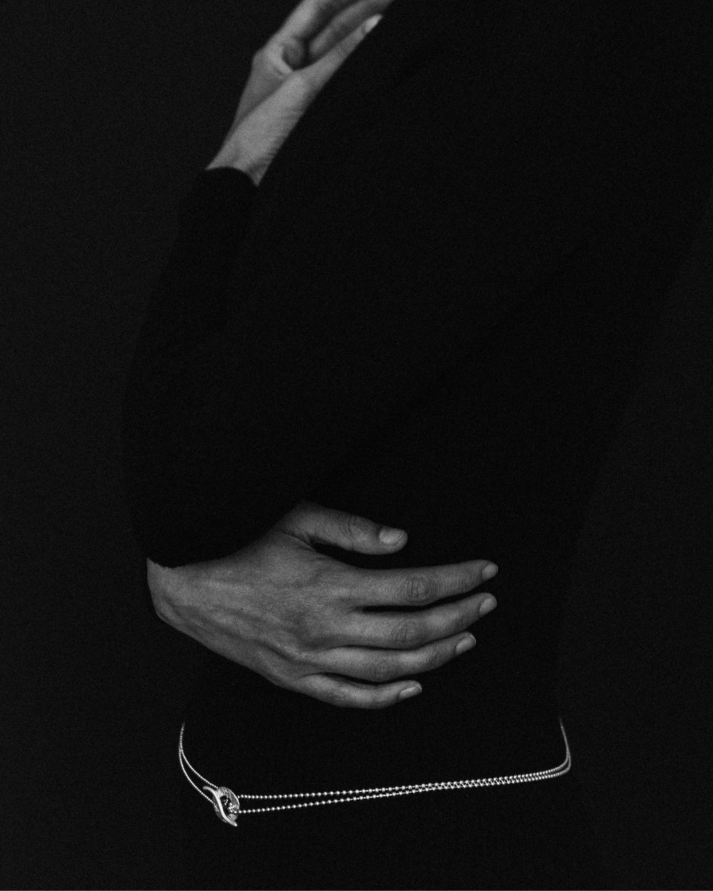 A black-and-white close-up of a person wearing a delicate chain necklace at the waist, with one hand resting on the hip against a dark background.