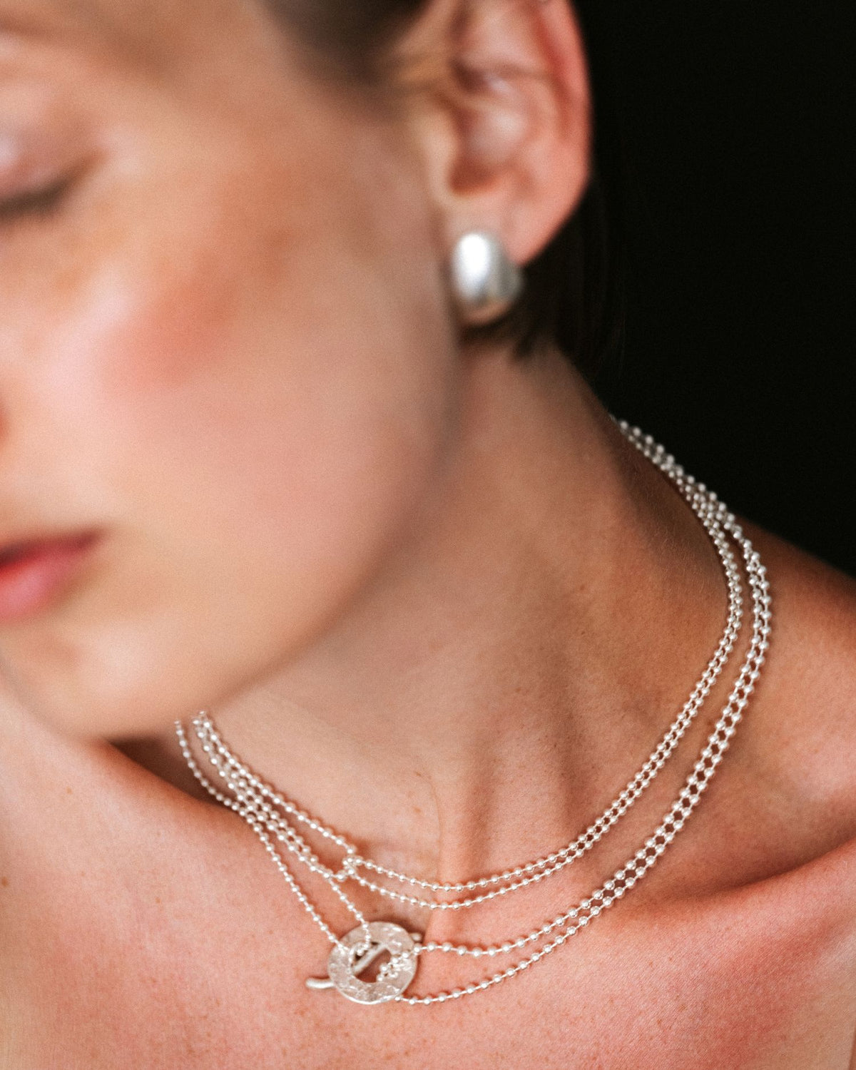 A close-up lifestyle shot of a model wearing a multi-strand silver necklace with a circular interlocking pendant and a silver stud earring against a dark background.