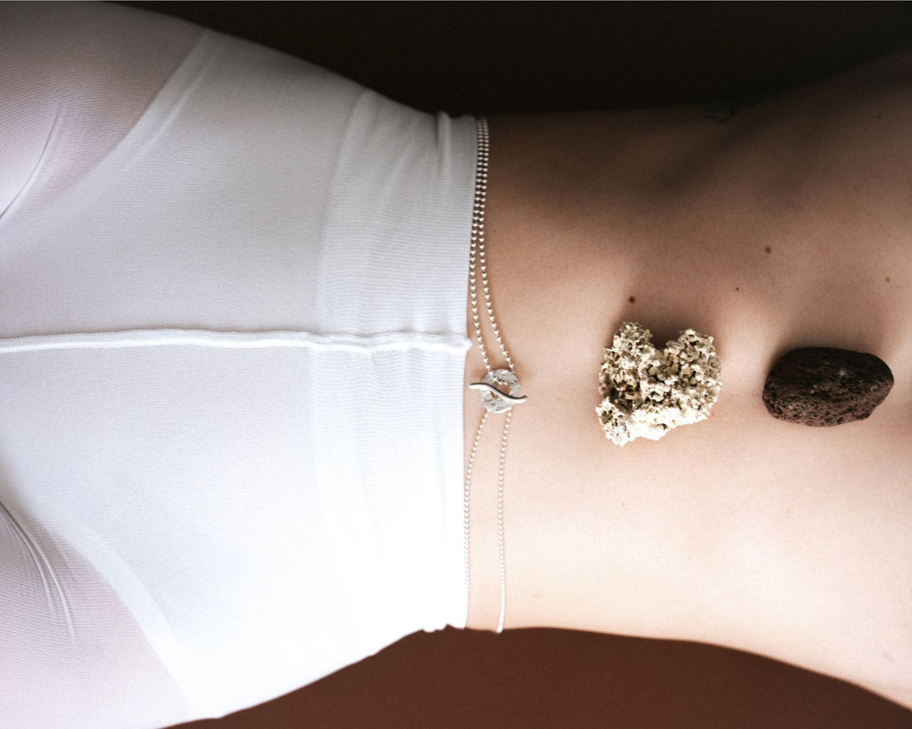 A close-up of a person's lower torso wearing white underwear and a delicate silver chain waist accessory, with two stones placed on the abdomen against a dark brown background.
