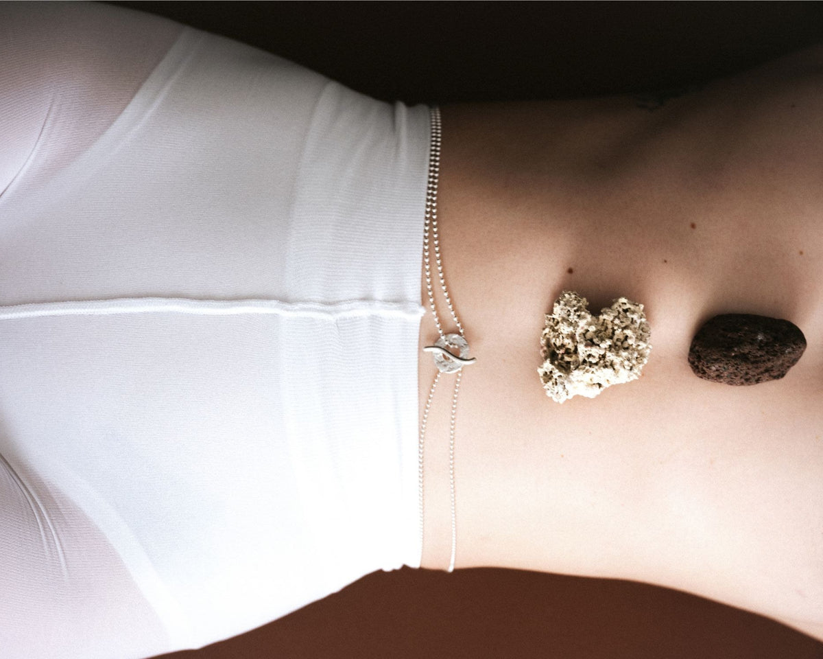 A close-up of a person's lower torso wearing white underwear and a delicate silver chain waist accessory, with two stones placed on the abdomen against a dark brown background.
