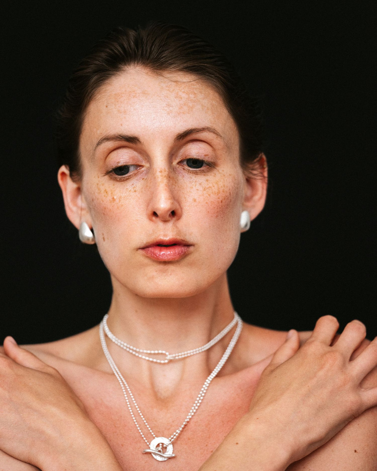 A close-up portrait of a woman with freckles wearing layered ball chain necklaces with t-bar clasp and matching silver stud earrings against a solid black background.