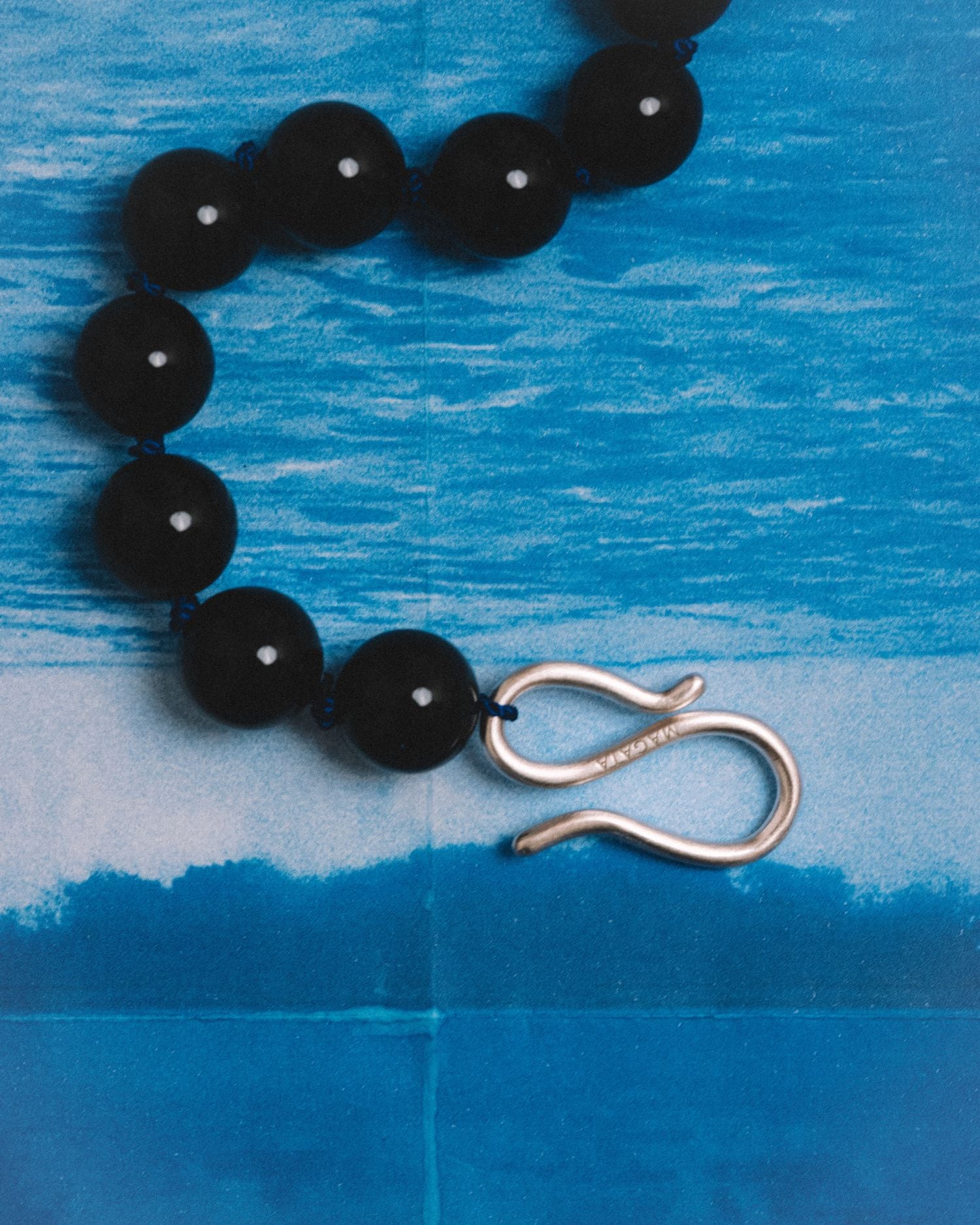 A close-up of a beaded necklace with glossy black round beads and a silver infinity-shaped clasp, laid on a blue textured background.
