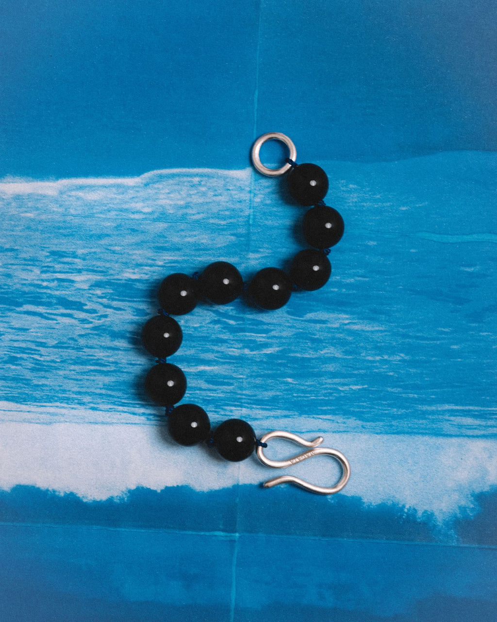 A beaded necklace with glossy black round beads and silver-toned metal hardware, including a lobster clasp and a circular ring, displayed against a blue ocean wave print background.