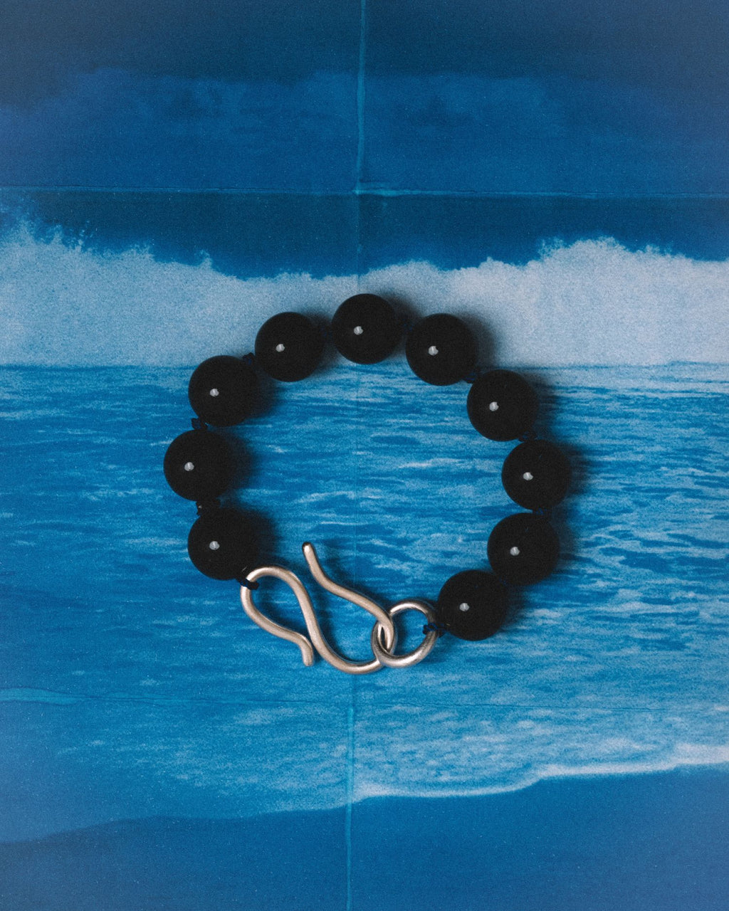 A black beaded bracelet with a polished silver infinity-shaped clasp, displayed against a blue ocean wave backdrop.