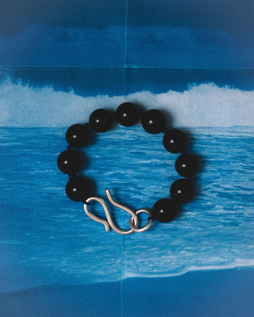 A black beaded bracelet with a polished silver infinity-shaped clasp, displayed against a blue ocean wave backdrop.