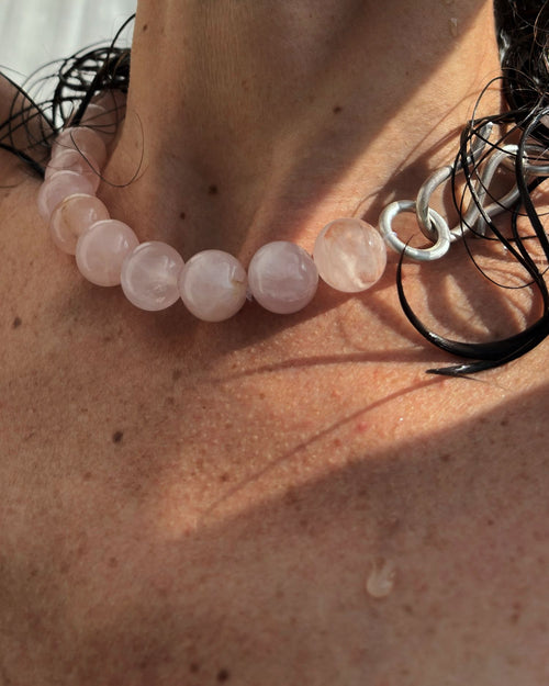 Close-up of a person wearing a beaded pink quartz necklace with a silver S hook clasp on a sunlit skin background.