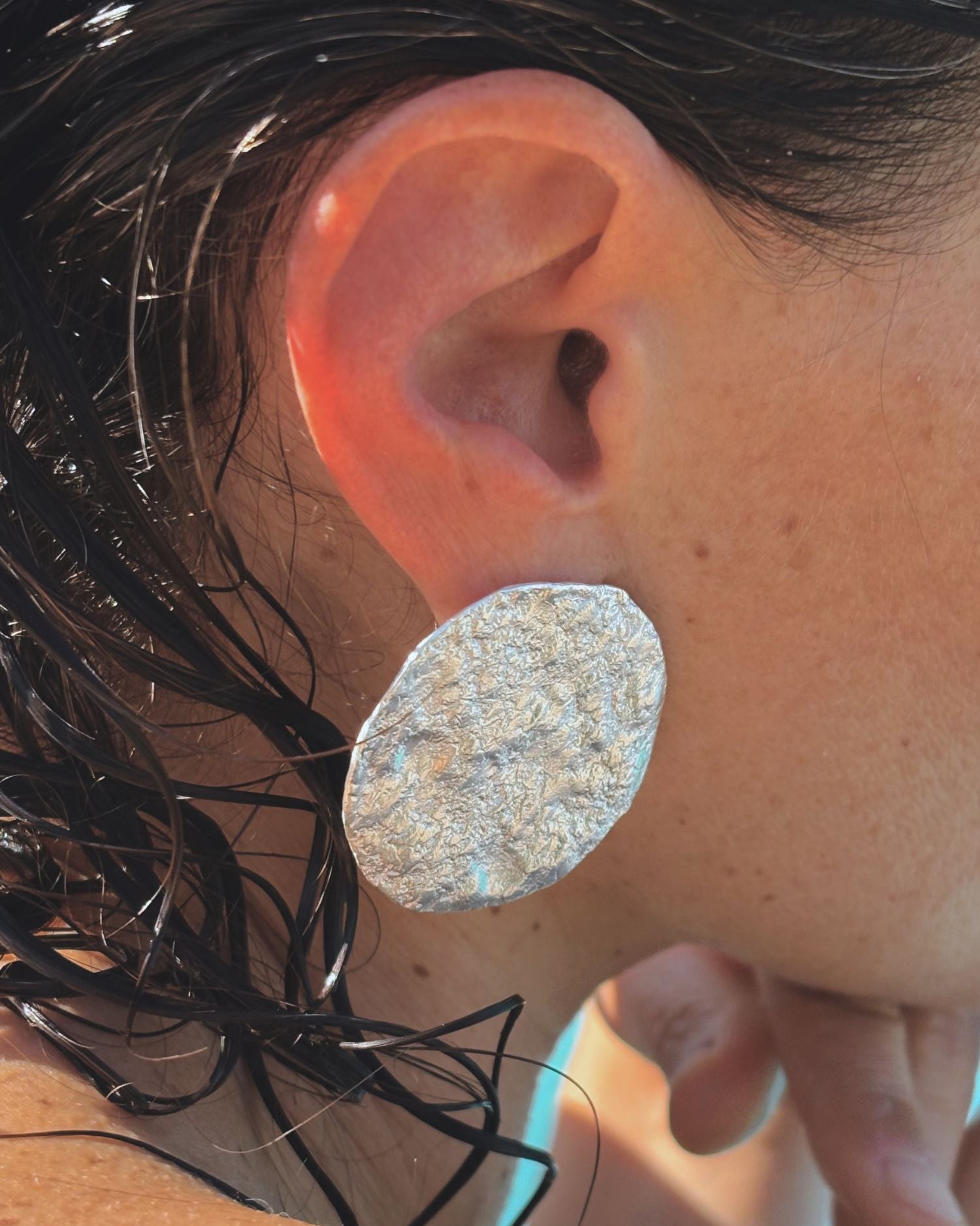 Close-up of an ear wearing a large, round, silver earring with moon textured surface.