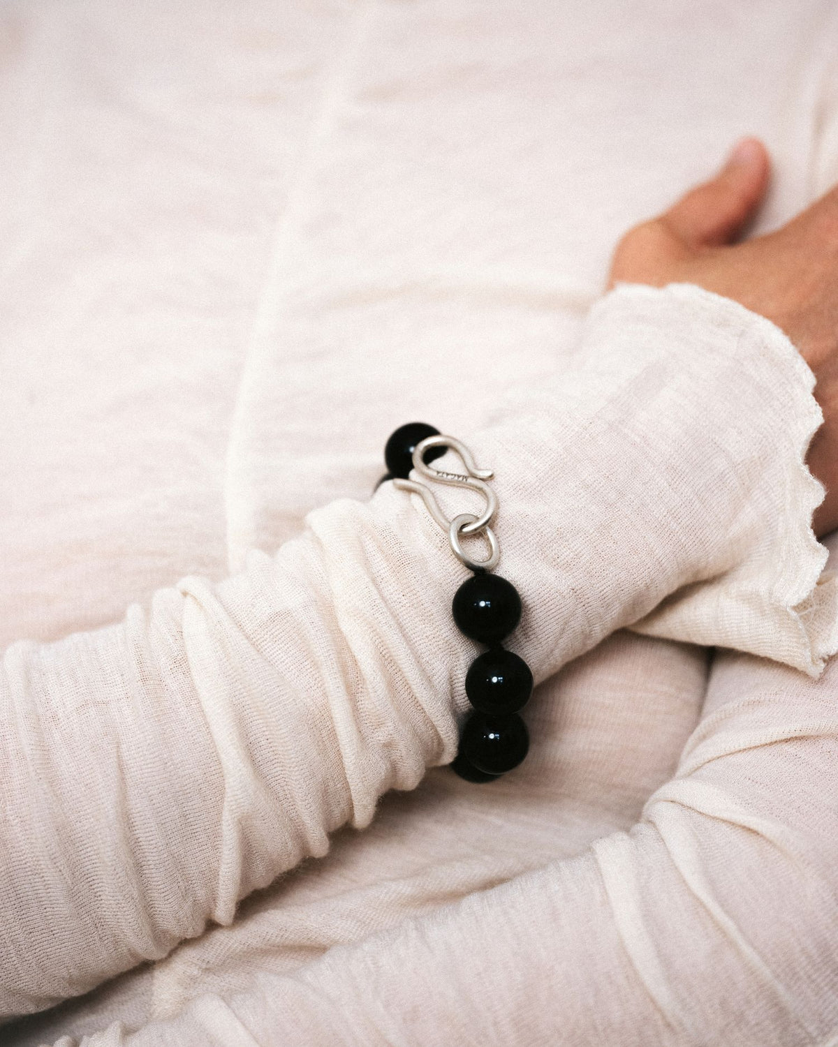 A close-up of a person's wrist wearing a black beaded onyx bracelet with a silver S-shaped clasp, styled with a white textured long-sleeve top.