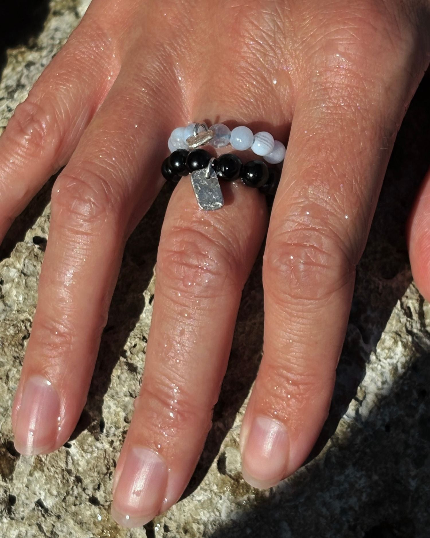A close-up of a hand wearing a beaded ring with black onyx beads, light blue chalcedony beads, and a small silver-toned geometric charm, resting on a textured stone surface.