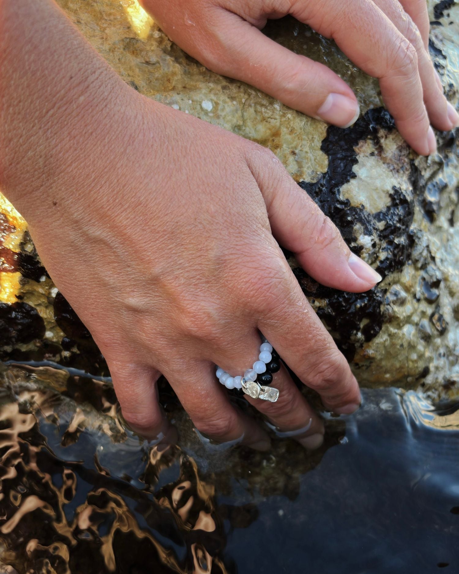 A close-up of a person's hand resting on wet rocks at the edge of shallow water, wearing a beaded chalcedony and black onyx ring with silver fragments.