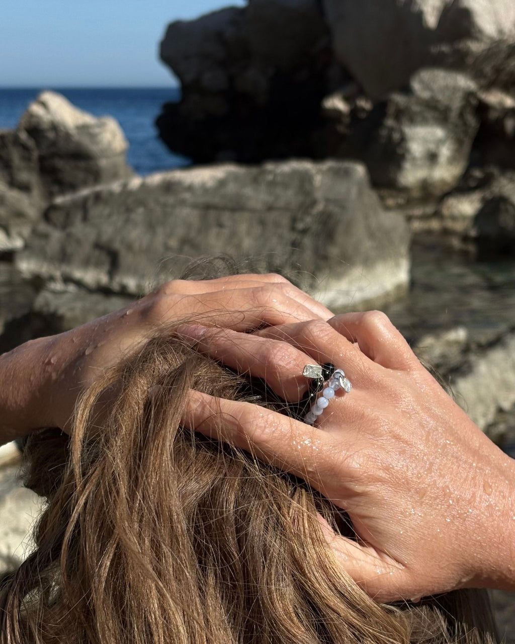 A close-up lifestyle shot of a person's hands holding wet brown hair, showcasing a silver beaded ring with a dark onyx on one finger, set against a rocky coastal background with the blue sea visible.