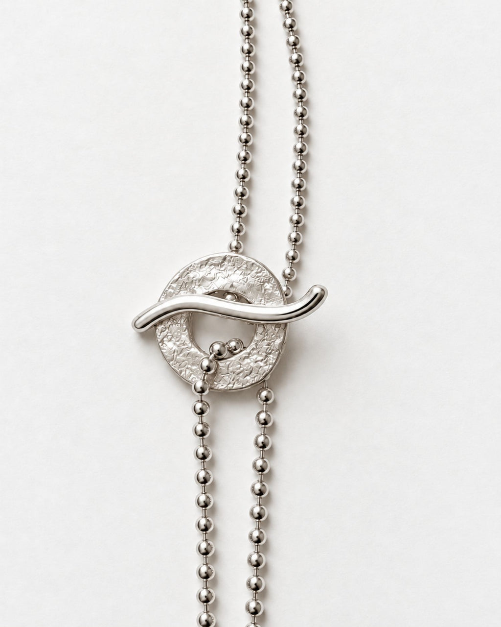 A silver ball-chain necklace featuring a textured circular clasp with a curved wave-like bar crossing it, suspended from a beaded chain on a plain light background.