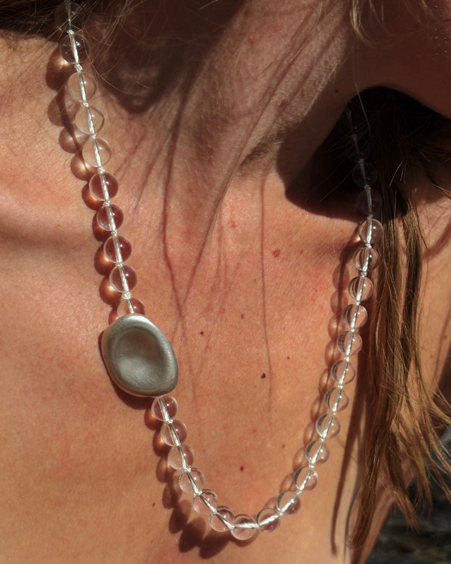 A close-up of a person wearing a beaded necklace with clear rock crystal beads, a silver-toned oval pendant, and a strand of translucent pinkish-brown beads.